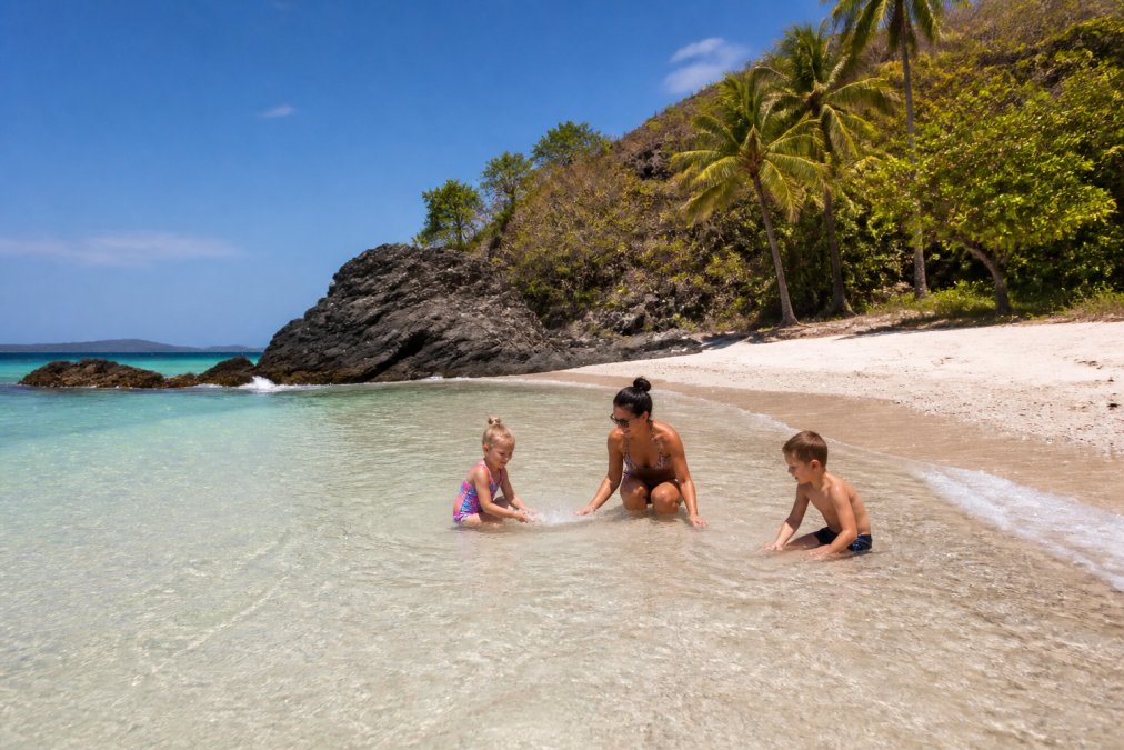 Family with young children playing in shallow, calm turquoise water at a secluded beach with white sand and rocky outcrops, palm trees in background, clear sky