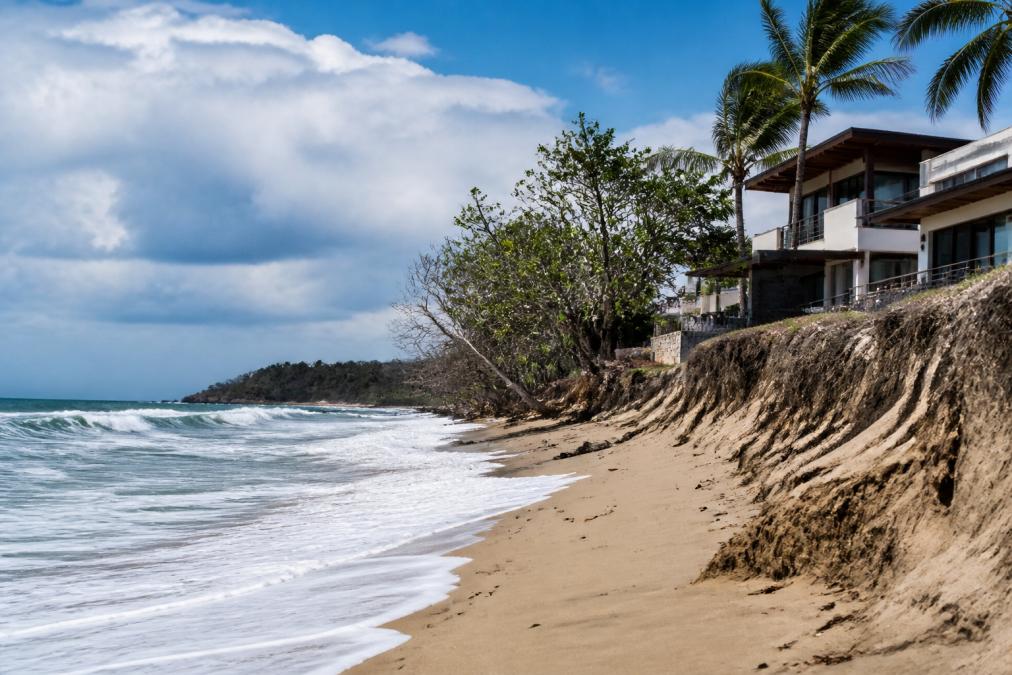 Coastal property in Guanacaste, Costa Rica with ocean views showing white-capped waves, sandy beach with visible erosion patterns, and modern luxury home perched near shoreline under partly cloudy sky
