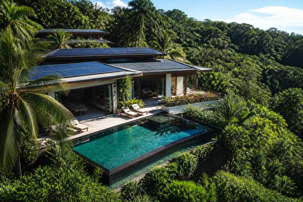 Aerial view of luxury villa with solar panels integrated into roofline, private infinity pool overlooking dense rainforest canopy, and lush native vegetation surrounding the property