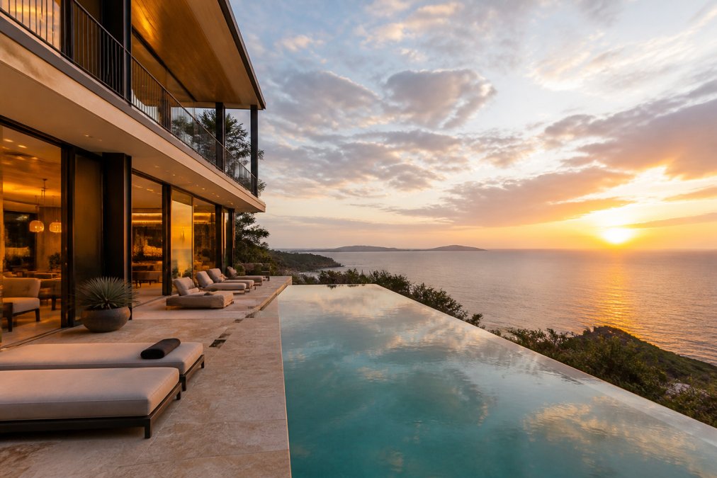 Stunning luxury oceanfront villa at sunset with illuminated terrace, infinity pool overlooking the ocean, modern furnishings visible through floor-to-ceiling windows, golden hour lighting