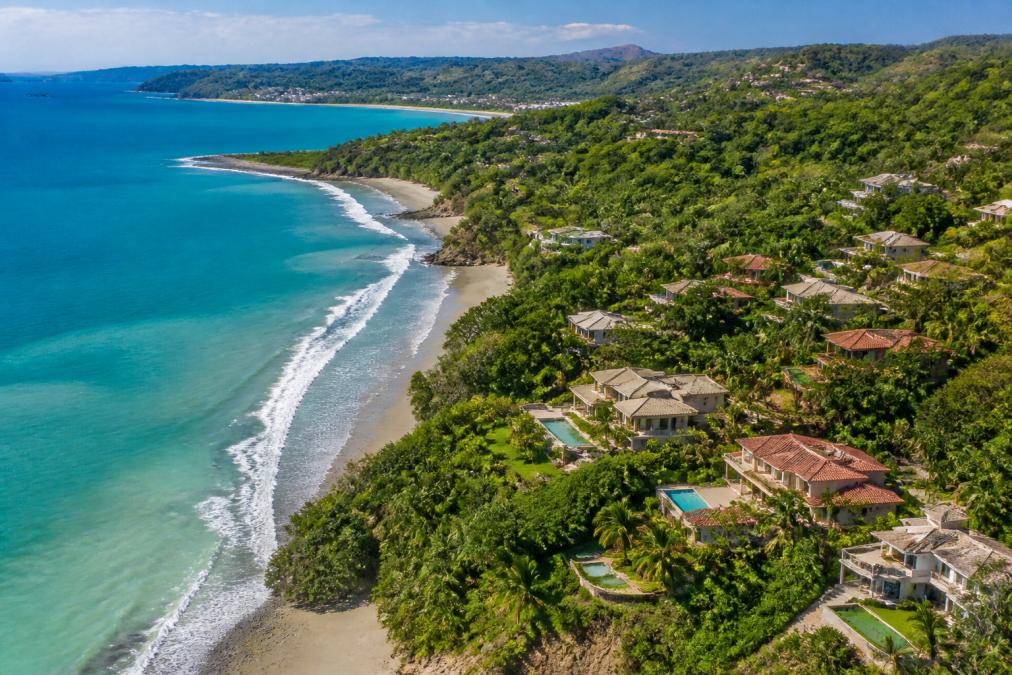 Aerial view of Tamarindo's beachfront properties with turquoise waters, palm-lined shores, and luxury homes nestled among tropical vegetation under bright afternoon sunlight