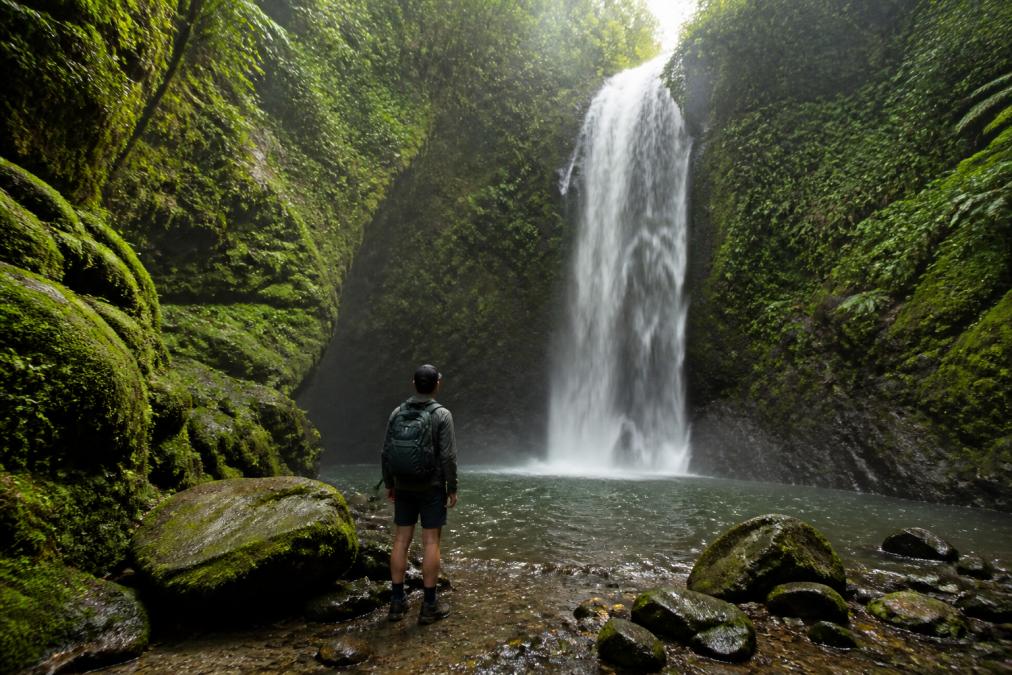 A solo hiker with a small backpack standing at the base of a wide jungle waterfall surrounded by moss-covered rocks and dense cloud forest vegetation in a remote Costa Rican national park, soft diffused light filtering through the canopy